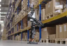 Humanoid Robots in Warehouse Environments – Trials Conducted Robotic warehouse worker standing on a concrete floor among tall pallet racks filled with cardboard boxes and crates.