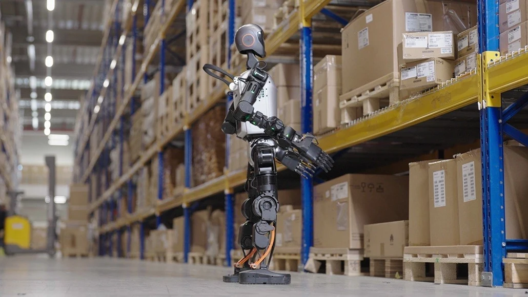 Robotic warehouse worker standing on a concrete floor among tall pallet racks filled with cardboard boxes and crates.