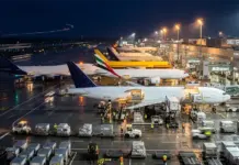 $1.2bn World Freight Acquisition by Brookfield on Cards Nighttime view of a busy cargo airport with several airplanes docked at gates and crates being loaded on the tarmac.
