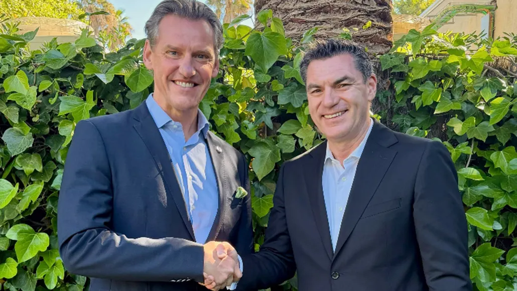 Two men in dark suits shake hands and smile outdoors in front of a green hedge.