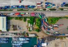 EcoRoute by DP World to Enhance Supply Chain Performance Aerial port scene with colorful shipping containers, trucks along a road, and a marina of boats at the bottom.