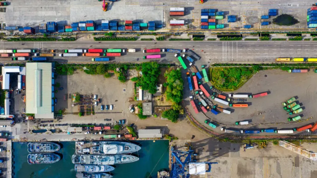 Aerial port scene with colorful shipping containers, trucks along a road, and a marina of boats at the bottom.