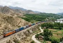 First International Freight Train Service Launched by China Orange freight locomotive pulling a long line of colorful shipping containers along a mountain railway beside a river valley.