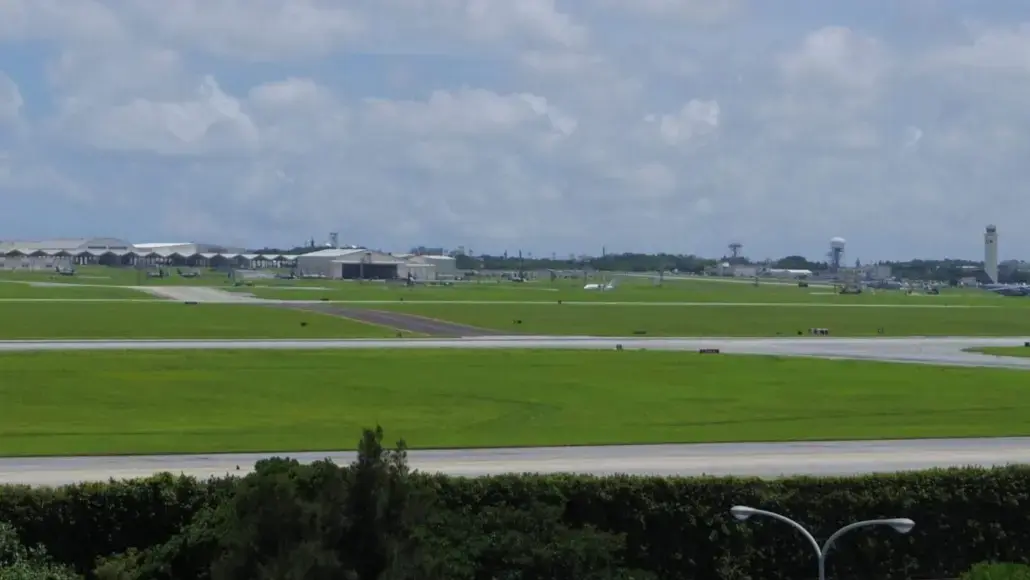Wide view of an airport with green grass fields, hangars, and a distant control tower under a cloudy sky.