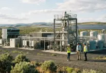 Supply Chain Diversification Through e-fuel Plant Launch Industrial energy site with large machinery and storage tanks in a rural landscape; four workers in safety vests and hard hats discuss the equipment (wind turbine visible in distance).