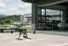 KMT Proposes Domestic Drone Procurement Budget of $7.52bn Quadcopter drone with camera on its landing gear on a tarmac, near a hangar with more drones and equipment.