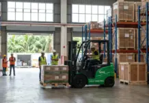Rhenus to Solidify its Warehousing Services in Philippines Green fully electric forklift lifting a pallet in a spacious warehouse as workers in safety vests and hard hats observe nearby by the loading bay.