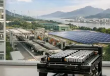CATL Aims Carbon Neutrality in Battery Value Chain by 2035 High-tech equipment rack with stacked modules in a bright indoor space, overlooking an outdoor energy plant with solar panels, wind turbines, and distant mountains across a river valley.